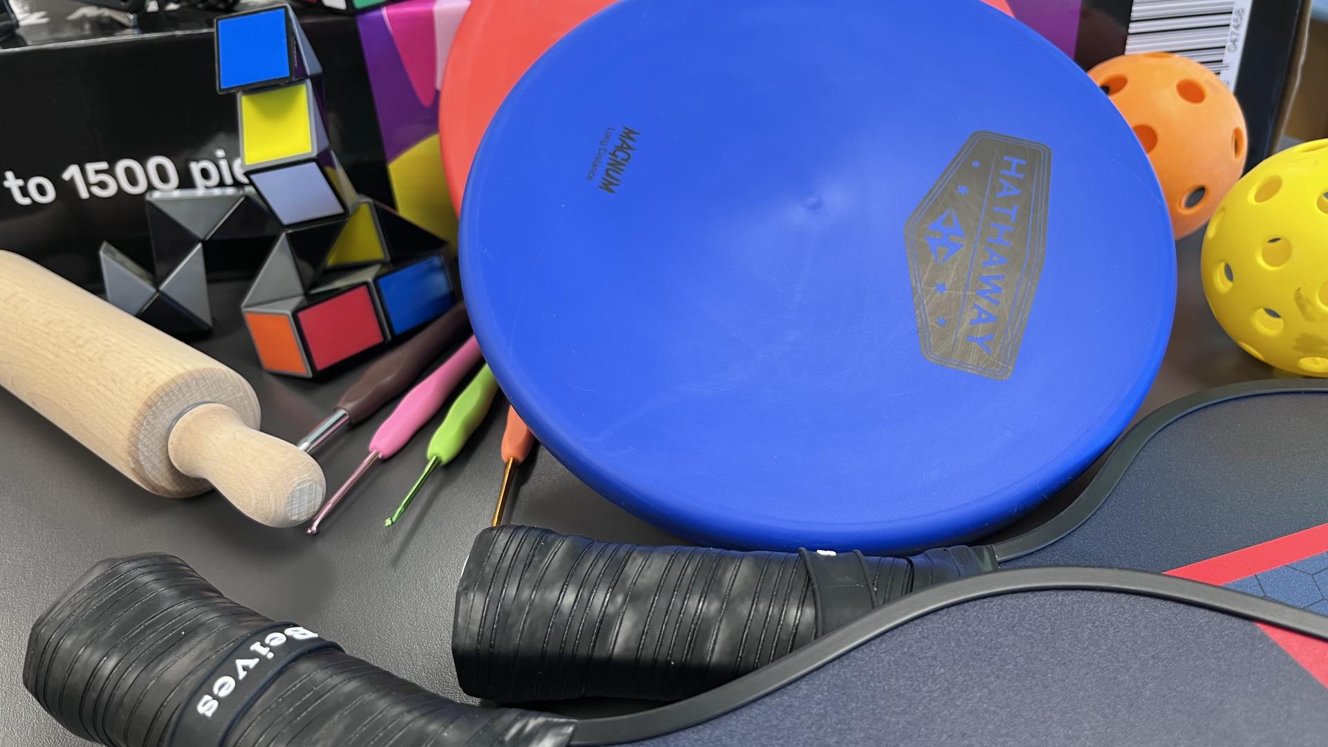 A close-up image of a collection of items, including, a rolling pin, disc golf discs, pickleball paddles and balls, Rubik's Cube, and crochet hooks.