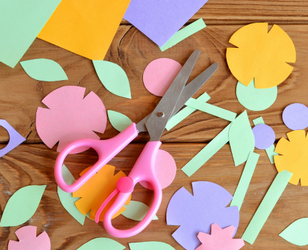 Pink handled children's scissors with paper flower pieces cutouts in yellow, pink and purple on a brown surface.